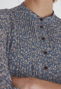 Vestido de estampado floral azul con cuello alto, diseño plisado y botones marrones. Presenta delicados patrones florales en blanco y rosa.