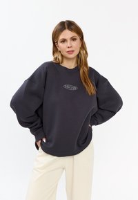Donkergrijze oversized sweatshirt met een ronde halslijn en een decoratieve logo in een lichtere tint in het midden. Zachte stof met geribde manchetten.