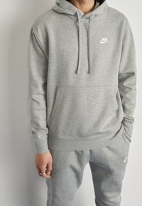Grå hoodie i mjukt material med känguruficka, dragsko på huvan och en liten vit Nike-logga på bröstet, kombinerad med matchande grå byxor.