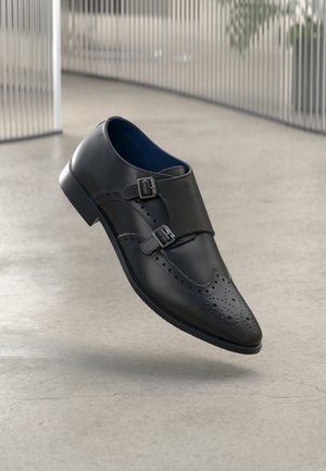 Chaussure monk strap en cuir noir avec double boucles et perforations décoratives, flottant au-dessus d'un sol lisse gris clair.