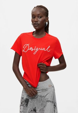 Donna con capelli intrecciati che indossa una maglietta rossa "Desigual" e una gonna grigia con stampa giornale, tenendo la maglietta leggermente sollevata.