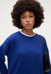 Felpa blu con scollatura tonda e vestibilità rilassata, caratterizzata da un colletto interno grigio a contrasto, abbinata a una borsa a spalla nera.