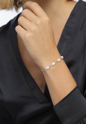 Vrouw met een zilveren armband met vijf witte parels, gekleed in een zwarte satijnen blouse, hand bij haar kin.
