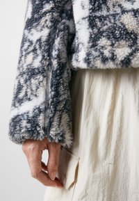 Fleece jacket in gray and white with a textured, plush surface. Features a zip closure and a hand resting on a cream-colored fabric.