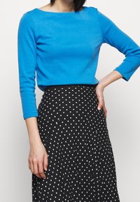 Blå långärmad topp med båturringning, som är ihop med en svart kjol med vita prickar och en plisserad design.