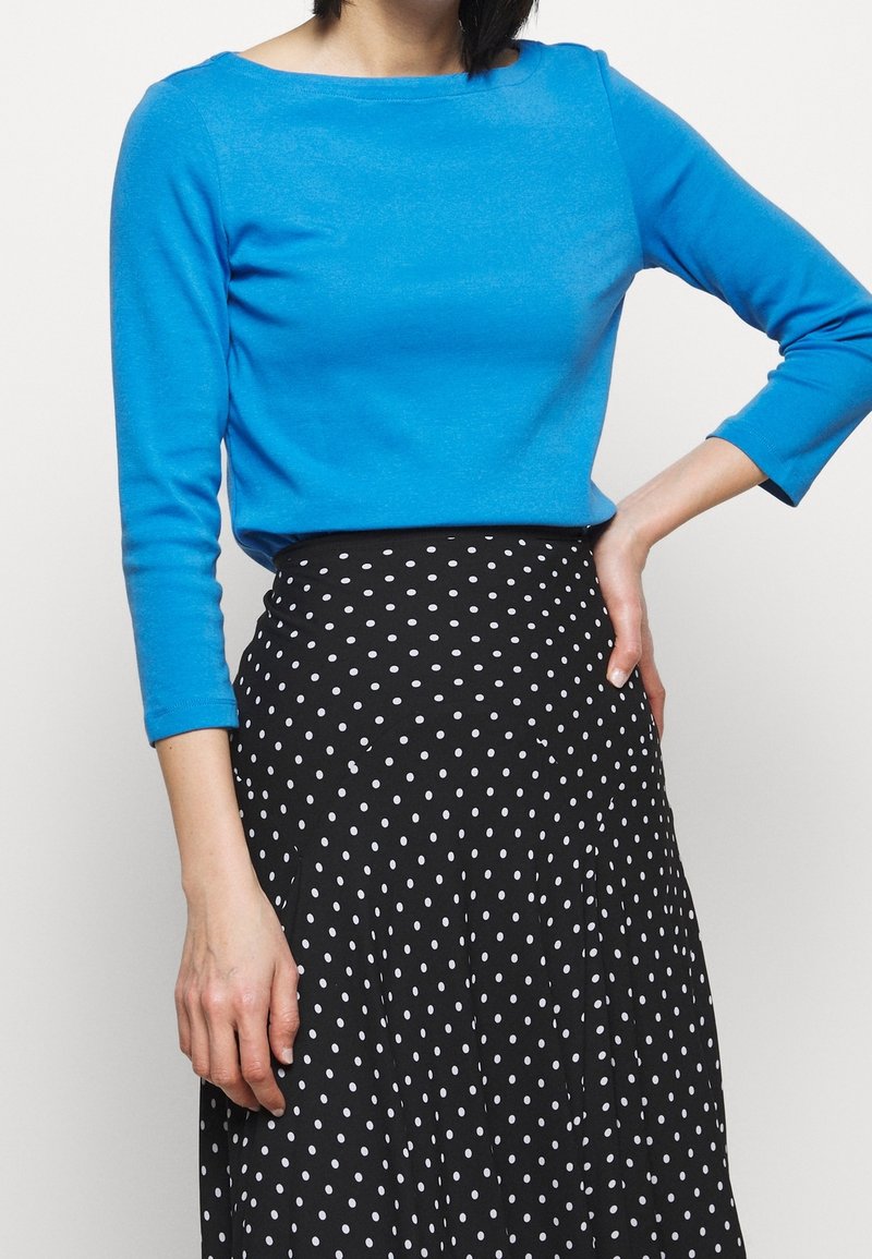 Blå långärmad topp med båturringning, som är ihop med en svart kjol med vita prickar och en plisserad design.