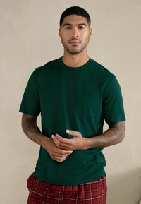 Maglietta verde scuro in cotone con scollo tondo e maniche corte. Presenta una tasca sul petto e una texture liscia. Indossata con pantaloni a quadri rossi.