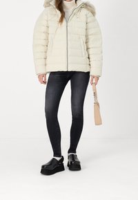 Cremefarvet puffjakke med hætte i imiteret pels, tekstureret quiltet design, lynlås foran, sorte skinny jeans og sorte platform-træsko med en beige clutch.