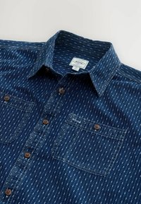 Knopfhemd aus dunkelblauem Denim mit einem Muster aus weißen Linien. Verfügt über zwei Fronttaschen und braune Knopfdetails.