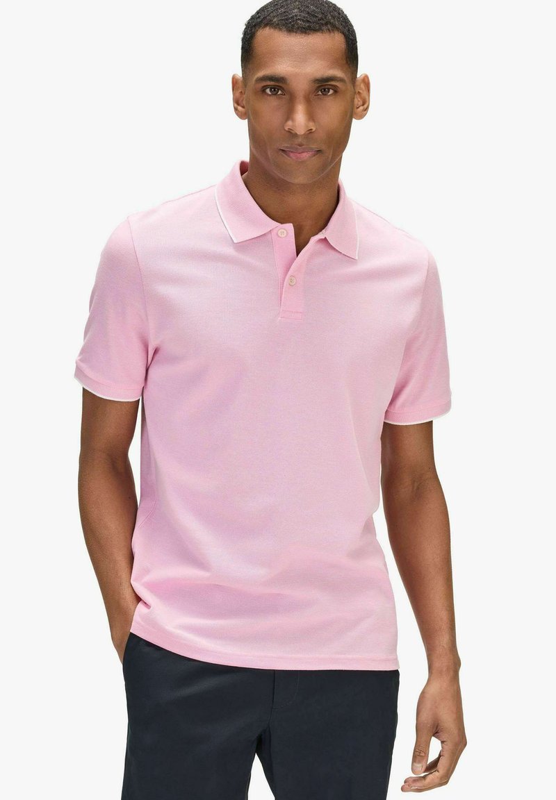 Homme portant un polo à manches courtes rose clair et un pantalon sombre, debout avec une main dans la poche contre un fond uni.