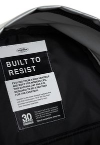 Interno di uno zaino Eastpak nero con etichetta in tessuto che mostra il testo in grassetto "PROGETTATO PER RESISTERE." Presenta materiali riciclati e una nota di garanzia.