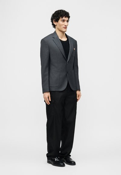 Jeune homme debout, portant un blazer gris ajusté sur une chemise noire, un pantalon noir ample et des chaussures élégantes noires, sur fond blanc uni.