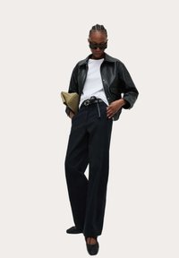 Zwart leren jasje over een witte shirt, hoge zwarte broek, zwarte ballerina's, houdt een gevoerde olijfgroene clutch vast, stijlvolle zonnebril.