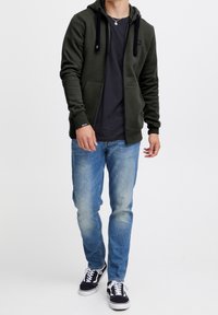 Felpa con zip verde scuro con accenti neri, tasche anteriori, indossata sopra una maglietta nera. Abbinata a jeans azzurri chiari e scarpe da ginnastica nere.