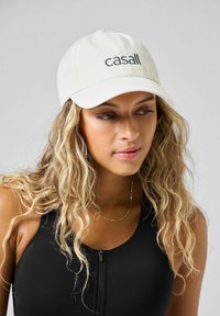 Biała czapka bejsbolowa z czarnym logo "casall". Wykonana z bawełny, z zakrzywionym daszkiem i sześcioma panelami. Modelka ma falowane blond włosy i nosi czarną bluzkę.