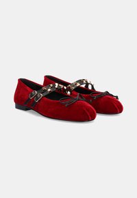 Bailarinas de terciopelo rojo con un tacón bajo, que cuentan con tiras negras entrecruzadas adornadas con detalles dorados y un lazo en la puntera.