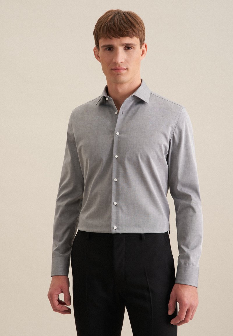 Camicia grigia a maniche lunghe con colletto, caratterizzata da una chiusura a bottoni, bottoni bianchi e una leggera texture. Indossata con pantaloni neri.