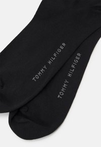 Schwarze Baumwollsocken mit dem eingewebten Text "TOMMY HILFIGER" in grau an der Seite. Glatte Textur, standardmäßiges Crew-Längen-Design.