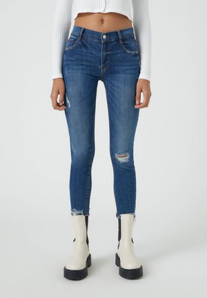 Vrouw draagt blauwe skinny jeans met hoge taille, distressed details en rafelige zomen, gecombineerd met off-white plateau laarzen tegen een effen achtergrond.