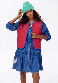 Prešívaná červená vesta s modrými akcentmi cez džínsové šaty s bielym vyšívaním a vzorom psa. Modelka nosí pastelovo zelenú čiapku.