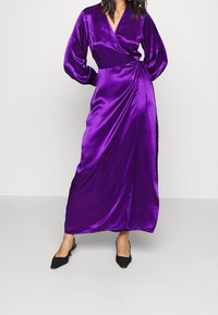 Lila Satin-Wickeldress mit langen Ärmeln, einem V-Ausschnitt und seitlichem Bindeband. Der Stoff hat eine glatte Textur und ein bodenlanges Design. Schwarze spitze Ballerinas.