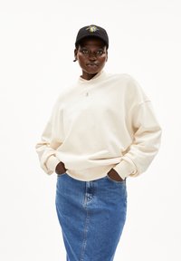 Crèmekleurige sweatshirt met een hoge hals, zachte textuur en subtiele logo. Gecombineerd met een lichtblauwe denimrok en een zwarte cap met een kleurrijk ontwerp.