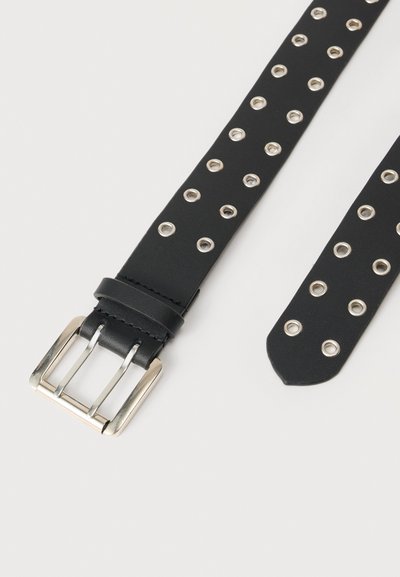 Μαύρη δερμάτινη ζώνη με ασημένια αγκράφα διπλού ακίδας και ομοιόμορφα τοποθετημένες ασημένιες οπές κατά μήκος της, πάνω σε λευκό φόντο.