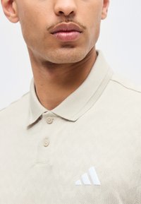 Κοντινό πλάνο άνδρα που φοράει μπεζ πόλο πουκάμισο με διακριτικό ζιγκ ζαγκ μοτίβο και λευκό λογότυπο Adidas στο στήθος.