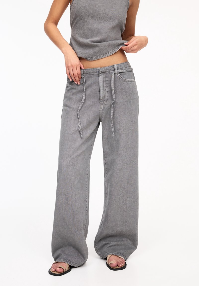 Personne portant un pantalon en jean gris ample avec des liens à l'avant, un top court gris sans manches et des sandales noires sur fond blanc.