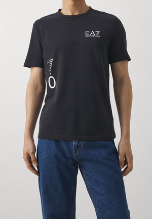 Černé tričko s krátkým rukávem s bílým logem "EA7 Emporio Armani" na hrudi a grafickým motivem na boku, nošené s modrými džíny.