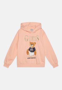 Ferskenfarget hettegenser med en grafikk av en bjørn i kjole med svart topp. "GUESS"-logo i gullbokstaver over illustrasjonen. Myk stoff.