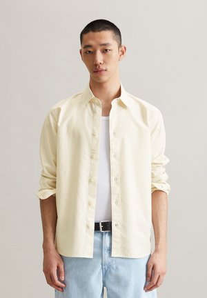 Light yellow striped button-up shirt with long sleeves, rolled at the cuffs, worn over a white tank top, paired with blue jeans.