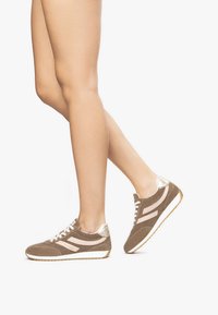 Baskets en daim marron avec accents métalliques argentés, présentant des bandes blanches sur les côtés, une semelle texturée et un design à lacets.