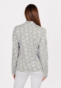 Donna con lunghi capelli castani che indossa un maglione grigio coperto da lettere bianche casuali, abbinato a pantaloni bianchi, di spalle rispetto alla fotocamera.