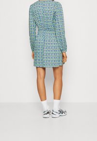 Vihreä kuviollinen mekko, jossa on pitkät hihat, joustava vyötärö ja lyhyt pituus, yhdistetty valkoisiin sukkiin ja lenkkareihin. Kuviot sisältävät sinisiä ja turkooseja yksityiskohtia.