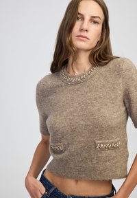 Kobieta w krótkim rękawie, puszystym brązowym swetrze z biżuteryjnym dekoltem i detalami kieszeni, zestawiona z niebieskimi dżinsami, na jednolitym tle.