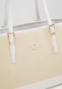 Sac à main structuré beige avec une surface tissée texturée, des accents en cuir blanc, quincaillerie dorée et un logo circulaire sur le devant.