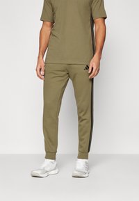 Olivenfarget treningstrøye med matchende joggebukser som har svarte sidestriper og Adidas-logo, kombinert med hvite sneakers.