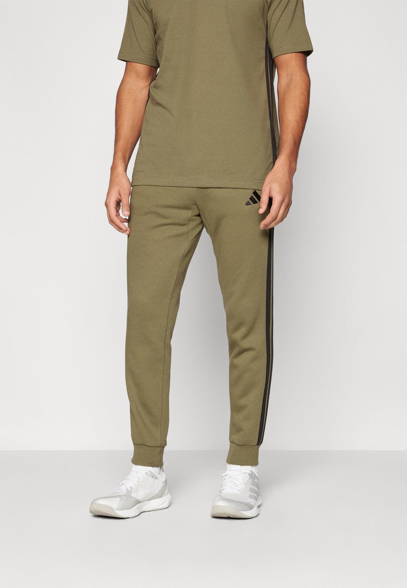 Olivenfarget treningstrøye med matchende joggebukser som har svarte sidestriper og Adidas-logo, kombinert med hvite sneakers.