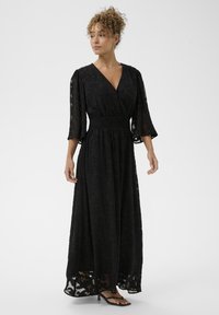 Fekete maxi ruha V-nyakkivágással, háromnegyedes ujjakkal és ráncolt derékkal. Áttetsző felső réteg texturált virágmintával. Szandálokkal viselve.