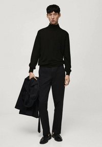 Svart polotröja, åtsittande stil, kombinerad med mörka byxor. Modellen håller en svart jacka och bär svarta skor. Enkel, strukturerad design.