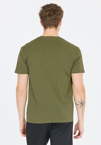 Olivengrøn T-shirt med korte ærmer, rund hals og en glat tekstur, der har en tatovering på højre underarm. Ingen synlige logoer.
