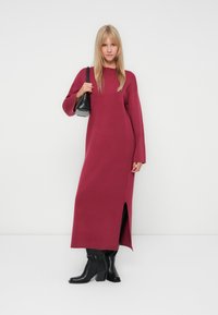 Abito a maglia bordeaux con maniche lunghe e spacco laterale, abbinato a stivali neri al ginocchio e a una piccola borsa nera. Texture liscia.