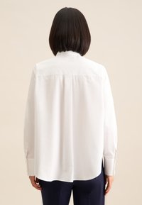 Camicia bianca a maniche lunghe con colletto alto, vestibilità rilassata e dettaglio piega sul retro. Presenta polsini con bottoni. Tessuto dalla texture liscia.