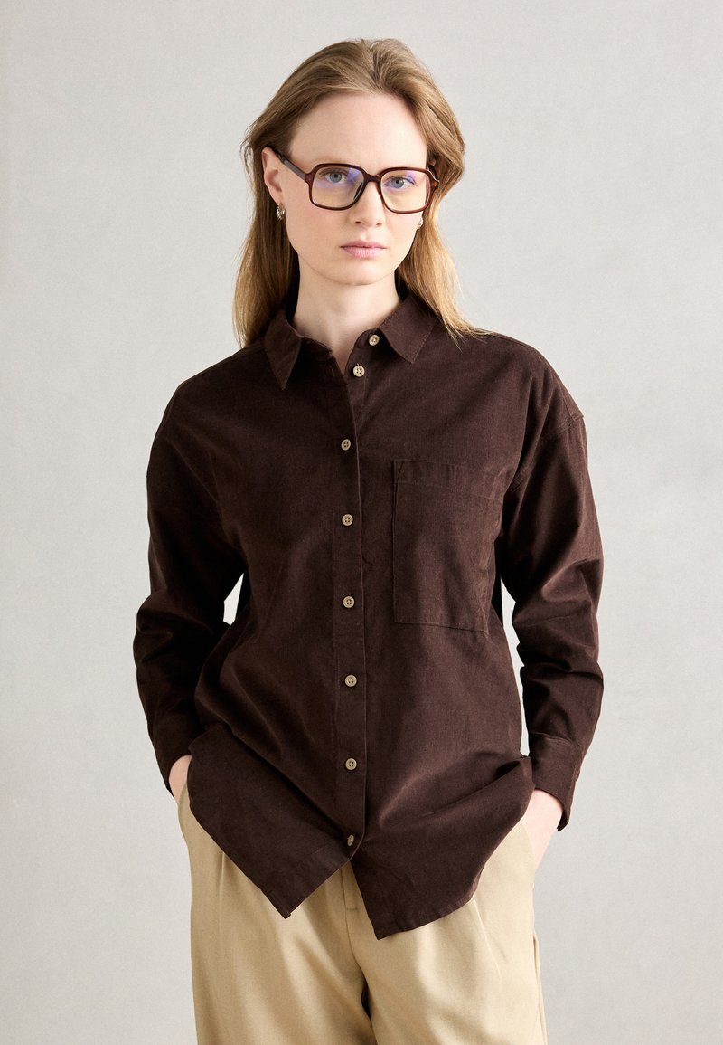 Brun skjorte med knapper i blødt stof med en brystlomme, der har en klassisk krave og brune knapper, parret med beige bukser.