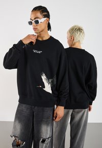 Black sweatshirt made of cotton with a graphic of a crane in flight. Features a crew neckline and loose fit. Grey distressed jeans worn.