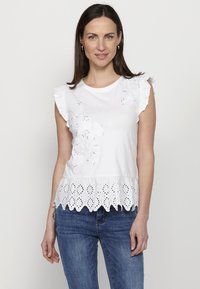 Top in cotone bianco con maniche a balze, ricamo floreale sul davanti e orlo in pizzo a occhiello con bordi ondulati. Indossato con jeans in denim blu.
