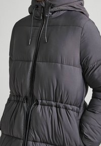 Šedá péřová bunda s kapucí, elastickým pasem a zapínáním na zip vpředu. Látka má hladký, lesklý povrch s prošitými detaily.