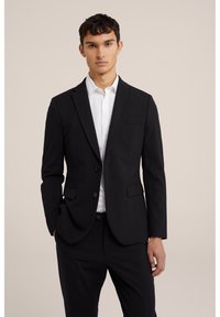 Blazer noir à la coupe slim et aux revers en notch, associé à une chemise blanche. Tissu lisse, fermeture à deux boutons, détail de poche avant.
