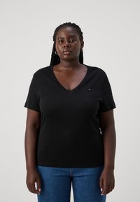 Camiseta negra de manga corta y escote en V, confeccionada con una tela suave, que presenta un pequeño logo en el pecho. Combinada con vaqueros azules.